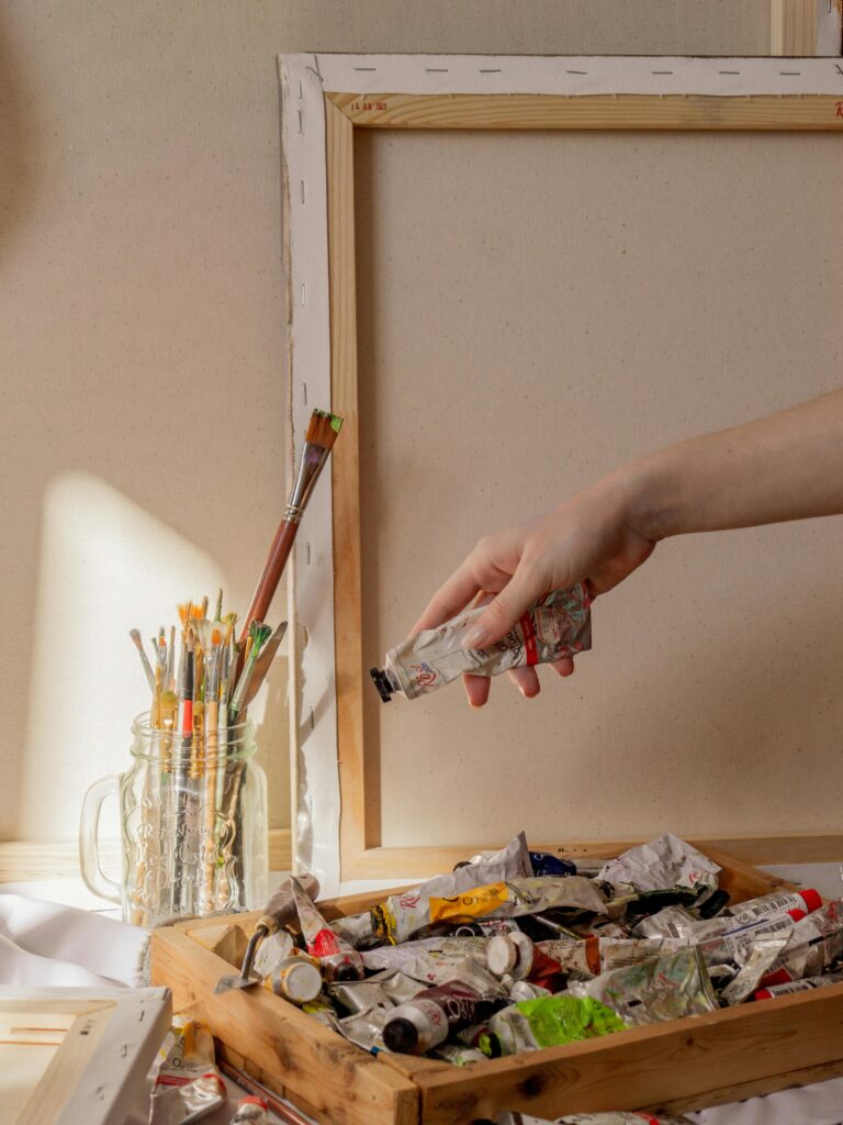 A hand reaching for paint tube in art studio with brushes and canvas frame.
